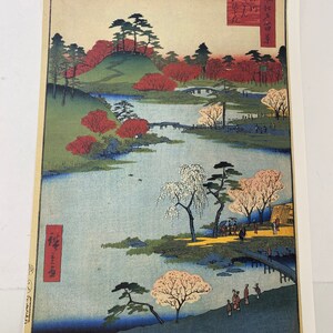 May include: A Japanese woodblock print depicting a serene landscape with a river winding through a lush green valley. The scene is filled with vibrant colors, including shades of red, green, blue, and white. The print features a bridge crossing the river, trees with blooming flowers, and a small village in the distance. The artist's signature is visible in the lower left corner.
