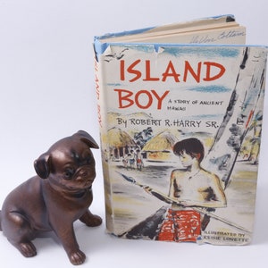 Könnte beinhalten: Ein Vintage-Hardcover-Buch mit dem Titel "Island Boy: A Story of Ancient Hawaii" von Robert R. Harry Sr. Der Buchumschlag zeigt eine Illustration eines jungen Jungen in einem roten Lendenschurz, der einen Speer hält und in einem Kanu steht, vor dem Hintergrund einer tropischen Inselszene.