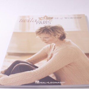 May include: A book cover featuring a woman in a light-colored sweater, with the text "twila paris" and "house of worship" visible. The book is published by Hal Leonard.