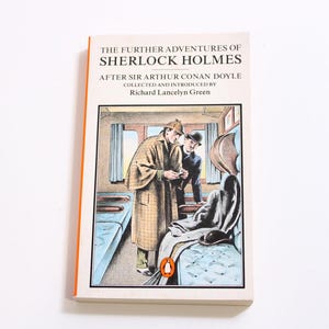May include: A paperback book titled "The Further Adventures of Sherlock Holmes" by Arthur Conan Doyle. The cover illustration depicts two men in a train car. The book has an orange spine and a Penguin logo.