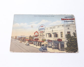 May include: A vintage postcard showing a street scene in a city. The postcard features a row of buildings with neon signs, including "Follies" and "Straight America Whiskey". There are cars parked on the road and people walking on the pavement.