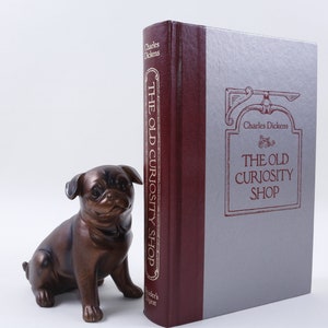 Op de afbeelding: Een bordeauxrood en zilveren hardcover boek getiteld "The Old Curiosity Shop" van Charles Dickens. Het boek ligt op een wit oppervlak naast een bronzen mopsbeeldje.