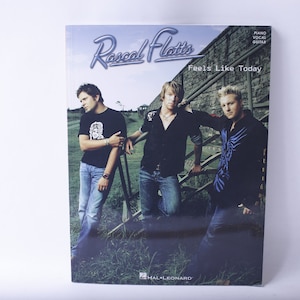 Può includere: Una copertina di un libro di spartiti con una foto del gruppo musicale country Rascal Flatts. La copertina è blu e presenta i membri della band in piedi davanti a un muro di pietra. Il titolo dell'album è "Feels Like Today".