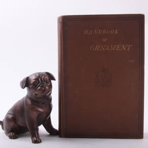 Könnte beinhalten: Eine bronzefarbene Mops-Figur steht neben einem großen, braunen Hardcover-Buch mit dem Titel "Handbook of Ornament". Das Buch hat goldene Schrift und ein dekoratives Emblem. Der Mops ist detailliert mit Falten und einer leichten Kopfneigung.