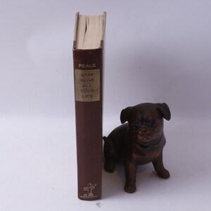 Peut inclure: Un livre relié en cuir marron intitulé "Stay Alive All Your Life" de Peale. Le livre est debout sur une surface blanche avec une petite figurine de chien en bronze devant lui.