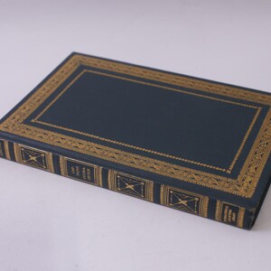 May include: A hardcover book with a dark blue cover and gold decorative trim. The book's spine features gold lettering and geometric designs. The book is lying flat on a white surface.
