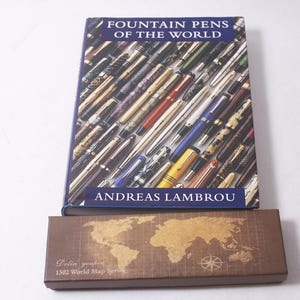 以下が含まれることがあります： 「FOUNTAIN PENS OF THE WORLD」というタイトルのアンドレアス・ランブロウ著の本。万年筆のコレクションが表紙に描かれています。本の下には、世界地図のデザインと「Dalin graphics 1502 World Map Series」の文字が入った茶色の箱があります。