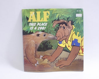 Può includere: Una copertina di un libro per bambini vintage con ALF, un personaggio alieno immaginario, con il testo "ALF This Place Is A Zoo!" La copertina presenta ALF con una camicia gialla con bordi blu, che guarda un formichiere marrone.
