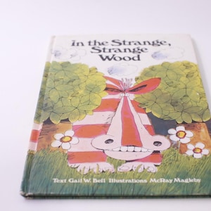 May include: A vintage children's book titled "In the Strange, Strange Wood." The cover features a whimsical illustration of a creature with red and white stripes, a large nose, and a wide grin, set against a backdrop of green foliage and white flowers.