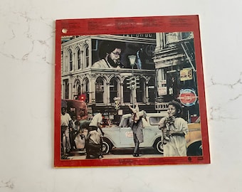 Può includere: Una copertina di un album di vinile vintage che presenta una scena di strada cittadina con un autobus rosso, un camioncino blu e persone che camminano. La copertina dell'album è rossa con un bordo giallo e ha il testo "The Rolling Stones" nella parte superiore.