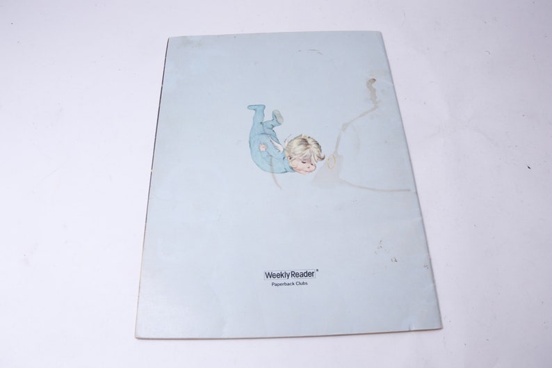 Pu&ograve; includere: Un libro tascabile vintage con una copertina azzurra che presenta un'illustrazione di un bambino in una tutina blu. Il testo "Weekly Reader Paperback Clubs" &egrave; stampato in basso sulla copertina.