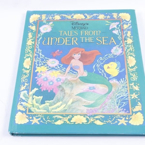 May include: A teal blue hardback book with a gold border. The book cover features an illustration of Ariel, the Little Mermaid, sitting on a rock with a yellow fish swimming nearby. The title of the book is "Disney's The Little Mermaid - Tales From Under The Sea."