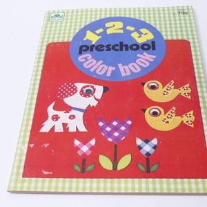 May include: A red cover with a green and white checkered border. The cover has a blue circle with the text "1-2-3 Preschool Color Book" in white. The cover also features a white dog with black spots, two yellow birds with black spots, and three flowers with checkered patterns.