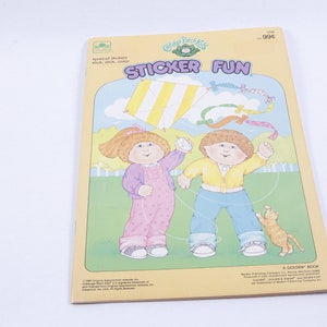 Puede incluir: Una cometa rayada de amarillo y blanco vuela en el cielo sobre dos muñecas Cabbage Patch Kids. Las muñecas están sujetando la cuerda de la cometa y sonriendo. La portada del libro dice "Sticker Fun" y presenta un logotipo verde de Cabbage Patch Kids.