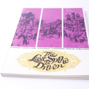 May include: A vintage book cover with a pink background and black and white illustrations. The title is "The Last Spike is Driven" and the text "National Golden Spike Centennial Commission Official Publication" is below the illustrations.