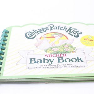 May include: A green and white Cabbage Patch Kids sticker book with a picture of a stork carrying a baby. The book has a spiral binding and says "Sticker Baby Book" and "16 Magic  Self-Stick  Stickers".