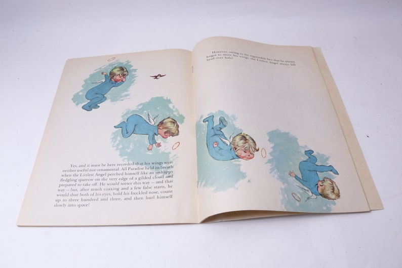 Pu&ograve; includere: Libro aperto con illustrazioni di un angioletto in pigiama blu con ali bianche, che cade nell'aria. Le illustrazioni sono in stile vintage, con testo sulle pagine.