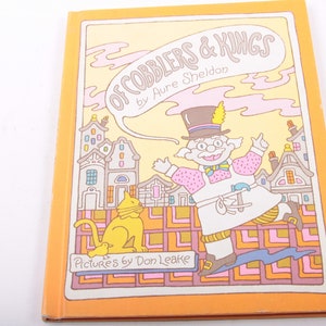 May include: A vintage children's book cover with an orange background. The title is "Of Cobblers & Kings" by Aure Sheldon. The illustration features a cartoon cobbler wearing a top hat and a cat.