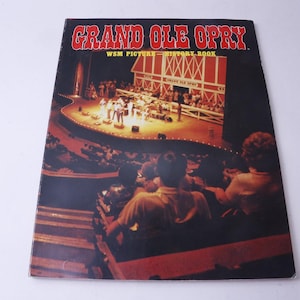 May include: A vintage "Grand Ole Opry" WSM Picture History Book. The cover features a stage scene with performers and a barn backdrop. The audience is visible in the foreground, with the title in red.