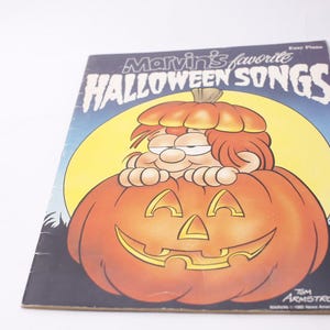 May include: A vintage songbook titled "Marvin's Favourite Halloween Songs." The cover shows a cartoon character peeking over a carved pumpkin with a smiling face, set against a yellow moon and a blue sky.