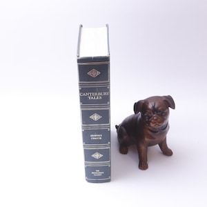 May include: A dark blue hardcover book with gold lettering titled "The Canterbury Tales" by Geoffrey Chaucer. The book is standing upright on a white surface next to a small brown ceramic dog figurine.