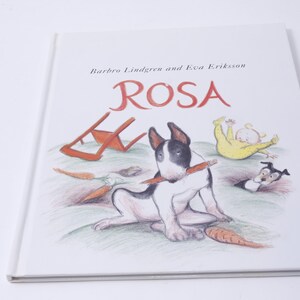 May include: A white and black bull terrier dog with a red pencil in its mouth. The dog is sitting on a green grassy surface with a red chair, carrots, and a small black and white dog in the background. The book cover reads "Barbro Lindgren and Eva Eriksson ROSA".