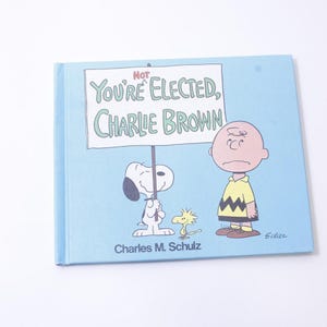 Pode incluir: Uma capa de livro azul com uma ilustração de desenho animado de Charlie Brown e Snoopy. O título do livro é "Not You're Elected, Charlie Brown".
