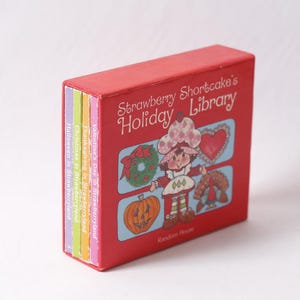 May include: A red box set labelled "Strawberry Shortcake's Holiday Library" featuring a cartoon Strawberry Shortcake character. The set includes small books with titles such as "Valentine's Day in Strawberryland" and "Halloween in Strawberryland."