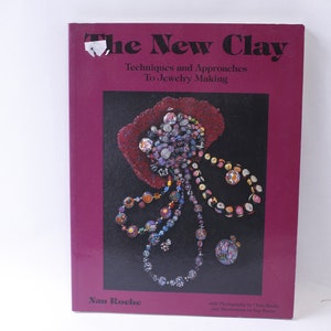 May include: A book titled "The New Clay" with the subtitle "Techniques and Approaches To Jewelry Making." The cover is a deep magenta color with a black background featuring a colorful clay jewelry design. The author's name is Nan Roche.