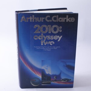 Pode incluir: Um livro azul de capa dura com um título prateado, "2010: odisseia dois" de Arthur C. Clarke. A capa apresenta uma ilustração azul e branca de uma nave espacial e uma citação de Carl Sagan.