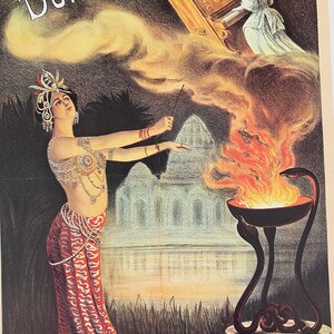 May include: A vintage poster advertising "DURGA" featuring a woman in a red and gold costume holding a wand, with a cloud of smoke behind her. A piano is floating in the air above her, and a fire burns in a bowl on a three-legged stand.