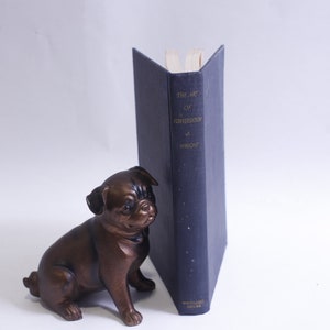 May include: A dark blue hardcover book titled "The Art of Conversation" by  J.  Wright. The book is standing upright on a white surface with a small bronze dog figurine in front of it.