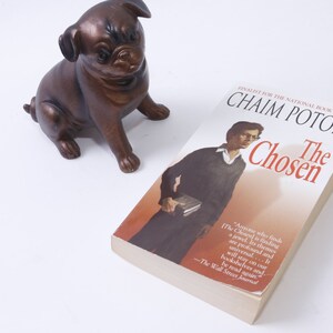 May include: A paperback copy of the book "The Chosen" by Chaim Potok. The book is a finalist for the National Book Award. The cover features a man wearing glasses and a dark sweater, holding a book. The background is a light orange color.