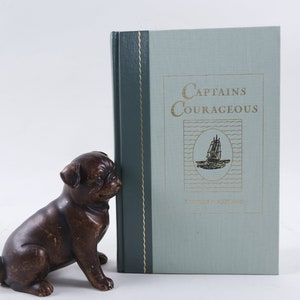 May include: A book titled "Captains Courageous" by Rudyard Kipling. The book cover is light blue with a gold illustration of a ship on the ocean. A brown pug figurine is next to the book.