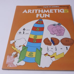 Puede incluir: Una portada de libro naranja brillante con el título "Arithmetic Fun" en letras grandes y en negrita. La portada presenta un cohete de dibujos animados con números en él, un cerdo rosa con monedas en la espalda y dos pasteles con arándanos. El libro forma parte de la serie Golden Fun at Home Workbooks.