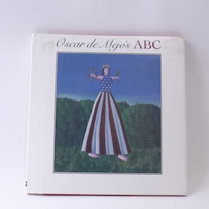 May include: A book cover with the title "Oscar de Mejo's ABC" in red lettering. The cover features a painting of a woman wearing a red, white, and blue dress with stars. She is holding a scale and a torch in each hand. The background is a blue sky with a crescent moon and a green forest.