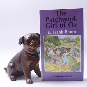 Może przedstawiać: Fioletowa książka w miękkiej oprawie zatytułowana "The Patchwork Girl of Oz" autorstwa L. Franka Bauma. Okładka przedstawia ilustrację trzech dzieci idących po żółtej drodze wybrukowanej.