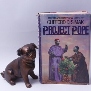 May include: A vintage paperback book titled "Project Pope" by Clifford D. Simak. The cover features a colorful illustration of two figures in a desert landscape.