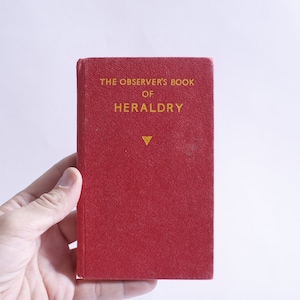 May include: A small, red book titled "THE OBSERVER'S BOOK OF HERALDRY" in gold lettering. The book has a textured cover and a gold emblem below the title. The book is held in a hand against a white background.