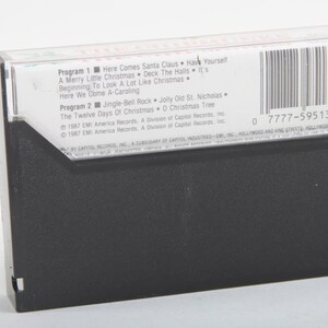 May include: A black cassette tape with a white label. The label lists the songs "Here Comes Santa Claus", "Have Yourself a Merry Little Christmas", "Deck the Halls", "It's Beginning to Look a Lot Like Christmas", "Here We Come A-Caroling", "Jingle Bell Rock", "Jolly Old St. Nicholas", "The Twelve Days of Christmas", and "O Christmas Tree". The label also includes the copyright information "1987 EMI America Records, A Division of Capitol Records, Inc.". The barcode is 0 7777-59513-4.