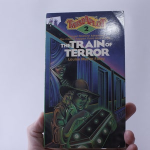 May include: A book cover for a mystery adventure novel titled "The Train of Terror" by Louise Munro Foley. The cover features a young girl in a scout uniform holding a box with glowing green lights. The background is a dark blue train car with a shadowy figure in the doorway.