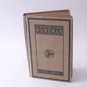 May include: A brown book with the title "The Manxman" by Hall Caine. The book cover features a green symbol of three legs.