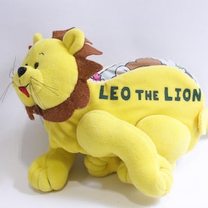 May include: A plush yellow lion toy with a brown mane and accents. The toy has the text "LEO THE LION" in green. The lion has a smiling face with whiskers and a red mouth. The toy is designed for children.