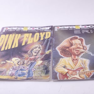 May include: Two vintage music magazines featuring illustrations of Pink Floyd and Eric Clapton. The Pink Floyd magazine has a yellow and purple cover. The Eric Clapton magazine has a black cover with a caricature of the musician.