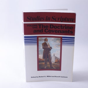 Può includere: Un libro bianco con una copertina rossa intitolato "Studies in Scripture, Volume One: The Doctrine and Covenants". La copertina presenta un'illustrazione in bianco e nero di un uomo in abito in piedi di fronte a un edificio.