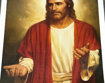 Op de afbeelding: Een ingelijst portret van een man met lang bruin haar en een baard, gekleed in een witte tuniek en een rode mantel. De man wordt afgebeeld met zijn handen open, tegen een zachte, gouden achtergrond.