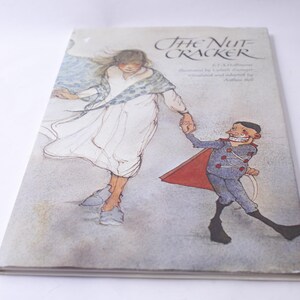 Peut inclure: Un exemplaire vintage du livre "The Nutcracker". La couverture présente une illustration d'une fille en robe blanche et d'un casse-noisette se tenant la main. Le titre est en écriture élégante, avec le nom de l'auteur et d'autres détails en dessous.