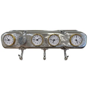 May include: A silver-toned metal automotive valve cover repurposed as a wall-mounted clock and hook rack. It features four round analog clocks with gold-colored bezels, labeled Los Angeles, New York, London, and Somewhere, with three spark plug hooks.