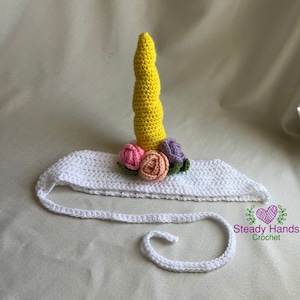 Puede incluir: Diadema blanca de ganchillo con un cuerno de unicornio amarillo de ganchillo y flores de ganchillo rosas, moradas y verdes. La diadema tiene una lazada blanca de ganchillo.