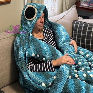 Puede incluir: Una manta de crochet azul y verde con un patrón de lunares blancos. La manta tiene una capucha grande de crochet con un parche de ojo negro. La manta está drapeada sobre una persona sentada en un sofá.